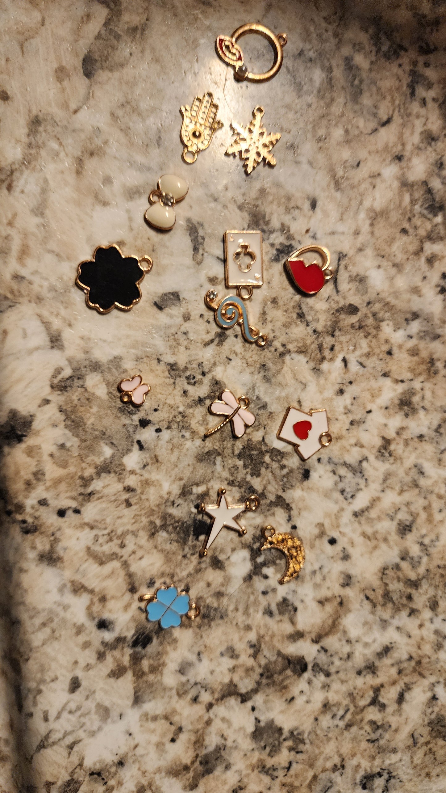 FAS Plant Charms: a collection of small gold and enamel charms—hamsa hand, hearts, flowers, crystals, crescent moon, dragonfly, key, and more—arranged on a speckled granite background.