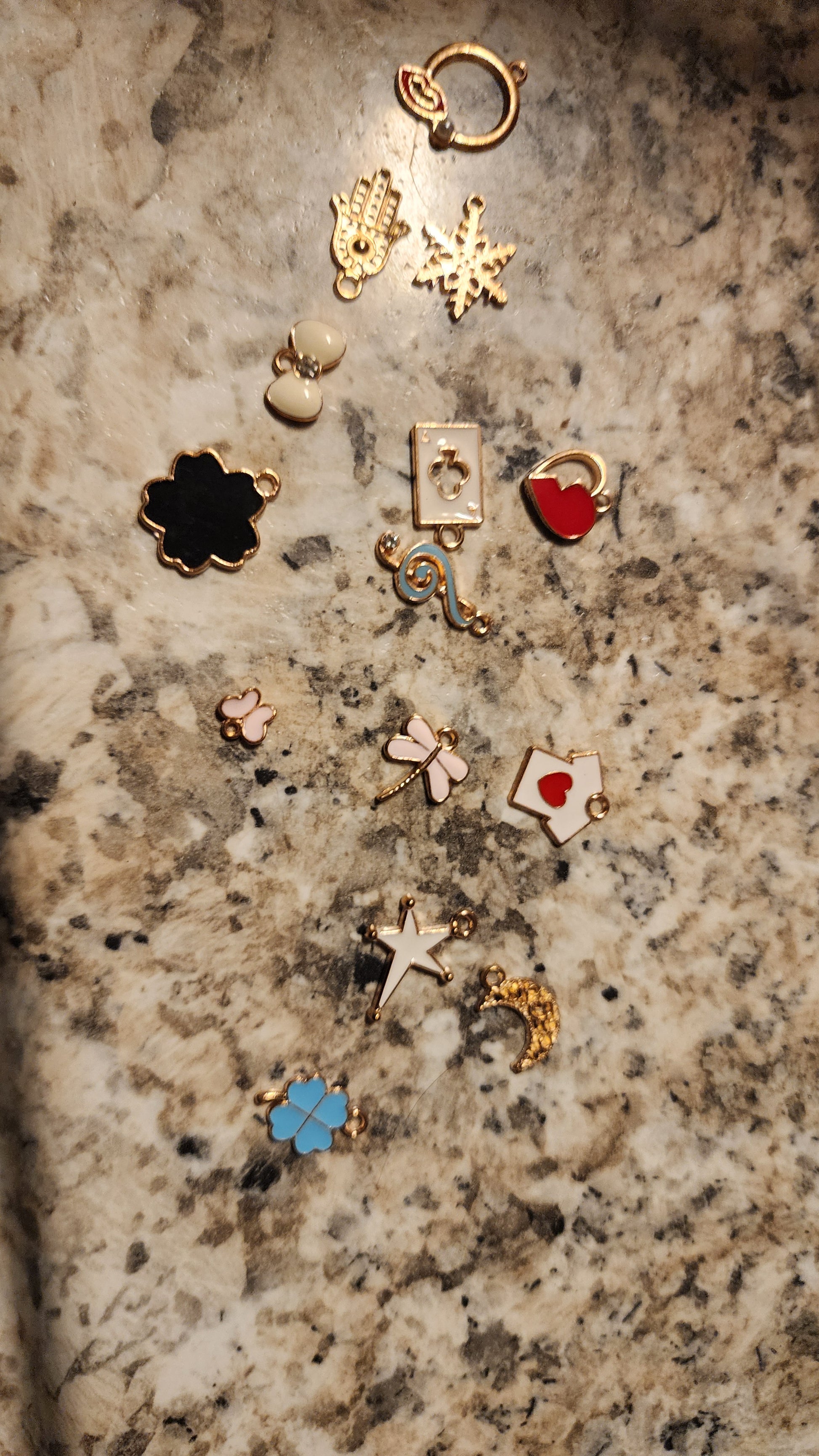 FAS Plant Charms: a collection of small gold and enamel charms—hamsa hand, hearts, flowers, crystals, crescent moon, dragonfly, key, and more—arranged on a speckled granite background.