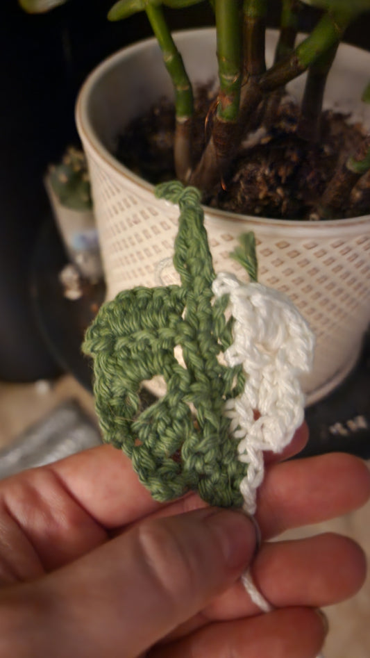 A hand displays a FAS Plant Charm, a small crocheted leaf in green and white, held in front of a potted plant with a textured cream pot on a polished wood stand.